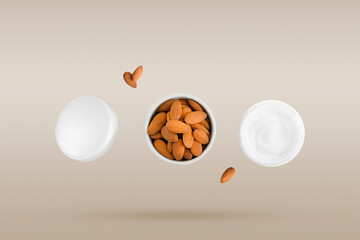 Opened round jar of cosmetic skin care cream and almonds in a ceramic bowl in a row falling on a beige background. Levitation concept. Natural ingredients for making cosmetics.