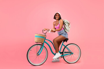 Full length portrait of lovely black woman in casual summer clothes riding bicycle over pink studio background