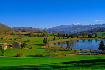Beautiful spring landscape with mountain lake and mountains covered with snow across blue sky. Fir trees, and trees reflected in water.  