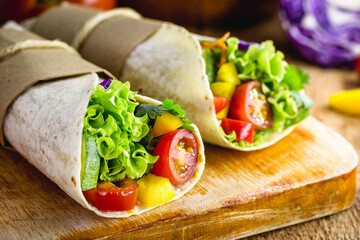 Two vegetarian tortilla wraps on wooden cutting board with vegetables in the background