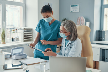 Beautiful young female nurse in protective mask talking to mature doctor while working in the...
