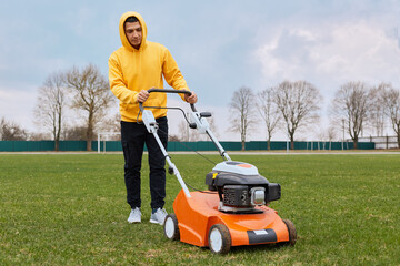 Full length photo of young adult male wearing yellow hoodie and black trousers, working in filed, mowing lawn with grass-cutter, looking at lawn mower with concentrated ans serious expression.