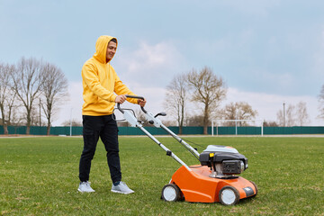 Handsome guy wearing casual closing working, mowing lawn with grass-cutter, gardener mows green grass with industrial gasoline mower, tends green, being in good mood.