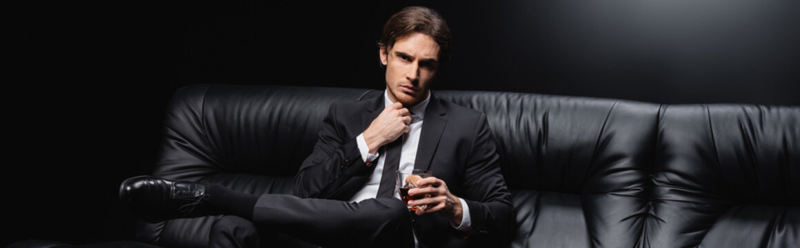 elegant man in suit sitting on leather couch with glass of whiskey on black background, banner.