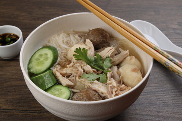 Clear white rice vermicelli beehoon noodle soup with chicken beef meat ball shredded cucumber in white bowl chopsticks spoon chili soya sauce parsley garnish