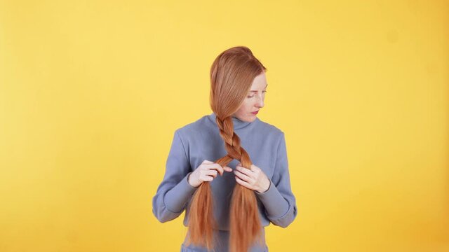 Attractive woman is making a plait from her beautiful long ginger hair. Hairstyle concept. Yellow background