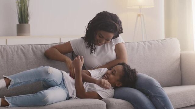 Little african american girl relaxing on mother's knees, young woman tickling her daughter, sitting on couch at home