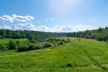 Naklejka na ściany i meble A green valley and a river in the distance under a blue sky with light clouds on a sunny day. Summer mood