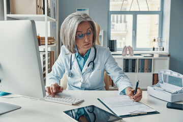 Serious mature female doctor in white lab coat writing something down while sitting in her office