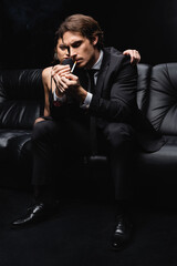 Fototapeta na wymiar woman in slip dress embracing man in suit holding lighter near cigarette on black.