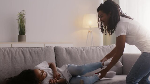 Happy african american mother and daughter playing together at home, woman catching girl and tickling her on sofa
