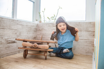 A boy with Down syndrome in a pilot's helmet sits on the floor near a large wooden model of an...