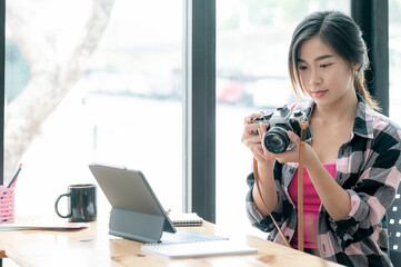 Young beautiful asian photographer holding and looking at camera screen while sitting at her studio.