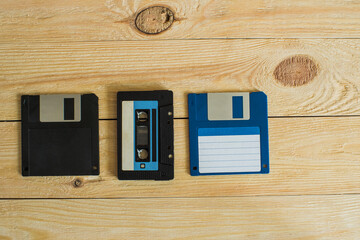 On a wooden background with knots is an audio cassette and two floppy disks. One is blue, the other...