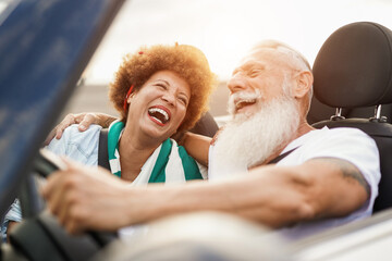 Trendy senior couple having fun inside convertible car - Multiracial mature people on a road trip in summer vacation with cabriolet car - obrazy, fototapety, plakaty