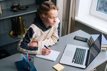 Top view of focused pupil boy listening teacher during online lesson using laptop computer via webcam video call. Child schoolboy doing homework at home. Concept of remote online distance education.