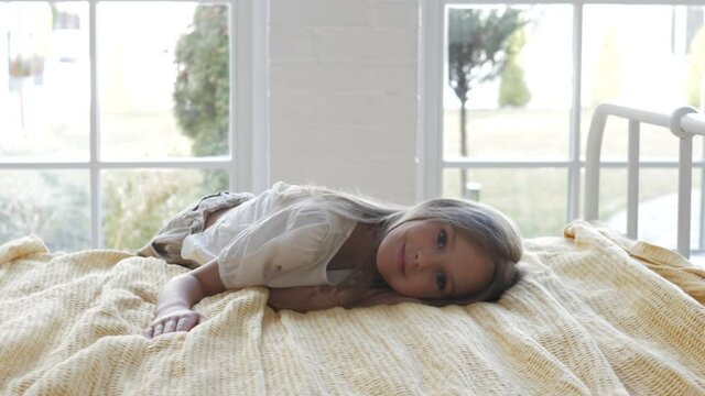 Adorable cute little female child kid girl lying on bed and looking at camera in the morning in cozy luxury bedroom in front of windows.