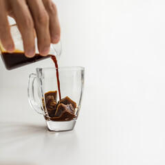 Hand pouring coffee from pitcher over black coffee ice cubes in transparent glass. White background, copy space, square crop. Summer refreshing beverage ice coffee concept.