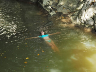 A girl in a blue swimsuit swims on her back in the muddy water of a rocky gorge in sun glare