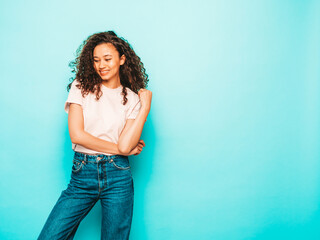 Beautiful black woman with afro curls hairstyle.Smiling model in white trendy t-shirt and jeans clothes. Sexy carefree female posing near blue in studio. Tanned and cheerful