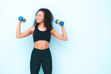 Fitness smiling black woman in sports clothing with afro curls hairstyle.She wearing sportswear. Young beautiful model with perfect tanned body.Female holding dumbbells in studio near light blue wall
