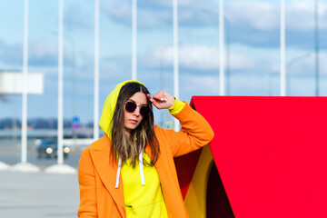 Millennial Hipster Girl in lilac sunglasses, orange coat, yellow hoodie posing at urban background. Trendy colorful casual outfit. Street Style. Fashion lookbook concept