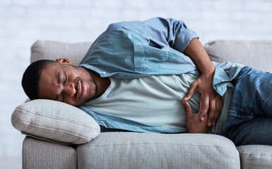 African Guy Suffering From Abdominal Pain Lying On Sofa Indoor