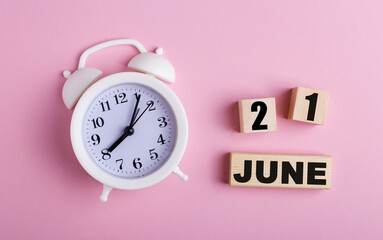 On a pink background, a white alarm clock and wooden cubes with the date of JUNE 21
