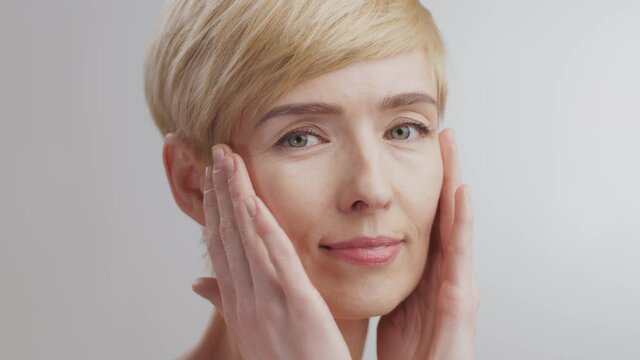 Profile portrait of middle aged woman massaging her face, applying pampering lotion, turning face to camera and smiling