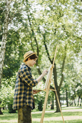 Naklejka na ściany i meble a young guy in a straw hat sits in the park in front of an easel and paints a picture with oil paints. Artistic work in nature. Plein air