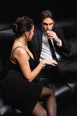 man in suit sitting on leather sofa with glass of whiskey near sexy woman in slip dress on black.