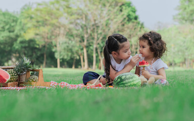 Two caucasian little cute girls sitting, playing in outdoor green park for picnic, eating fruit,...