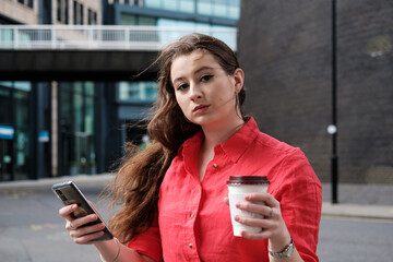 Young woman in a red shirt drinking a coffee and using her smartphone in the street.