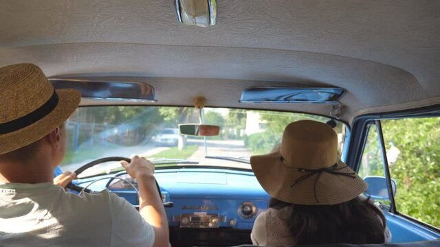 Young couple in hats driving on country road in classic vintage car. Unrecognizable pair riding into old auto. Man and woman sitting at the front seat of a retro automobile. Trip concept. Slow motion