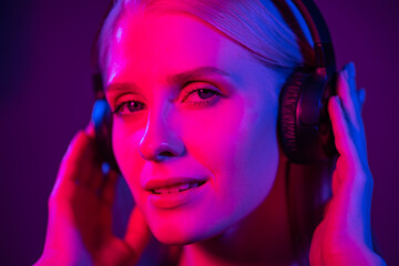 Young blonde woman listens to music on headphones at home with colored light. The girl smiles and looks into the camera. Close-up, blue and red lights. Home party, fun and entertainment concept.