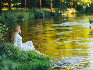 Cute beautiful young woman in white dress sitting on the river bank, sunny summer evening