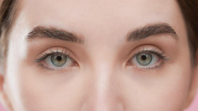 Extreme close-up of good-looking European female skincare model with brown hair opens her eyes moving her head up and looking at the camera against pink background | Skincare commercial
