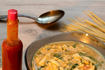 Spicy asian noodle soup in a glass bowl, tabasco sauce and uncooked noodle served on a wooden board 