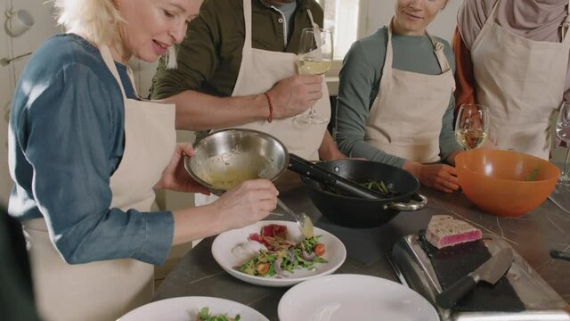High-angle medium shot with slowmo of woman serving dish on plate sprinkling it with oil and other people drinking wine and watching during cooking class with chef