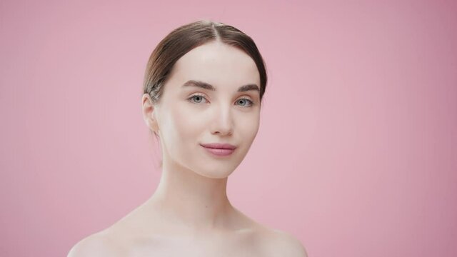 Attractive slim young European woman with dark hair and naked shoulders touches her cheek with cotton pad looking at the camera against pink ripple background | Eye cream commercial