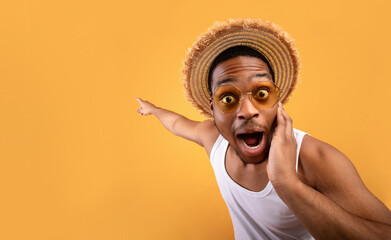 Summer sale. Shocked black guy in straw hat pointing at empty space on yelow studio background