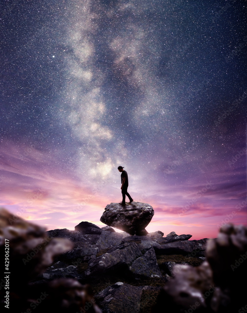 Wall mural a man standing on a rock as the milky way galaxy rises into the night sky at dusk. photo composite
