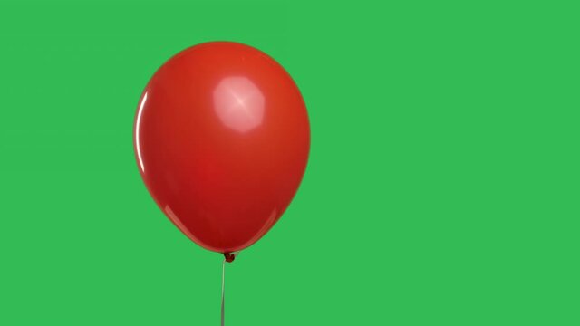 Red oval shaped balloon hanging in the air against the background of a green screen chroma key. Close up of colorful helium balloon. Birthday, party decoration, holiday, surprise, gift. Slow motion.