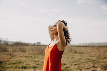 Young woman with curly hair, wearing a red summer dress, enjoying good weather, outdoors, in a...
