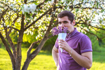 Defocus portrait of handsome caucasian man with cone flowers in lilac garden Funny enjoyment young brunette guy smelling fresh flowers and looking at camera on blurred background Out of focus