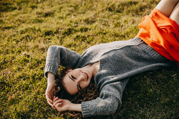 Young happy woman with curly hair, lying on grass in a field, wearing a sweater, smiling.