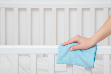 Young adult woman hand holding blue dry rag and wiping edge of white crib in bedroom. Closeup. Side...