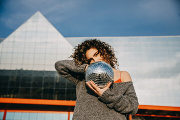Portrait of a young woman with curly hair, holding a disco ball, outdoors, on city background.