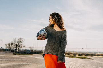 Young brunette woman with curly hair, walking with a disco ball, outdoors, in golden hour.