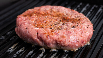 Hamburger patty getting cooked in a cast iron grill pan. The different seasonings is clearly visible on top of the patty.
Hamburger meat and food preparation concept.